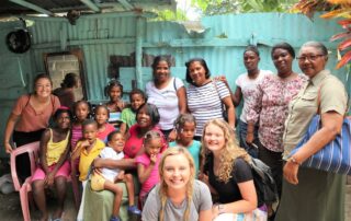 A group photo of Joni and Friends interns posing with families from the Dominican Republic. Many of the people in the photo are young children and their mothers or grandmothers. They appear to be in a tenement shack.