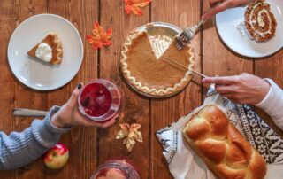 A scene from above a table with a pumpkin pie, a loaf of bread and fall leaves on it as someone is slicing the pie and another person is drinking a cranberry beverage.