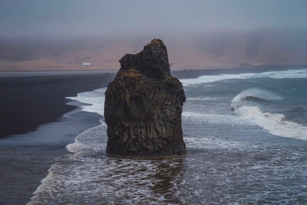 A large, standing rock in the middle of a shoreline, the waves crashing on it.