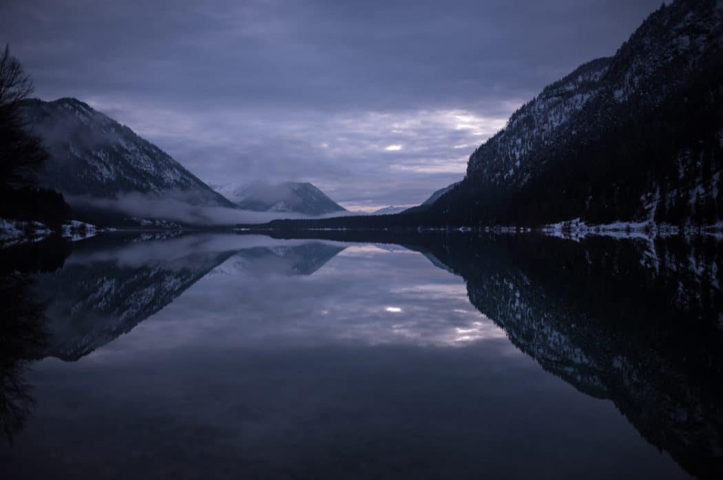 A body of water surrounded by dark, forested mountains, stormy clouds overhead.