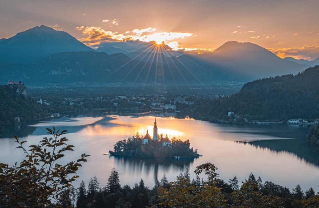 A picture of a castle on an island in the center of a lake with the sun rising over the mountain range in front of it.