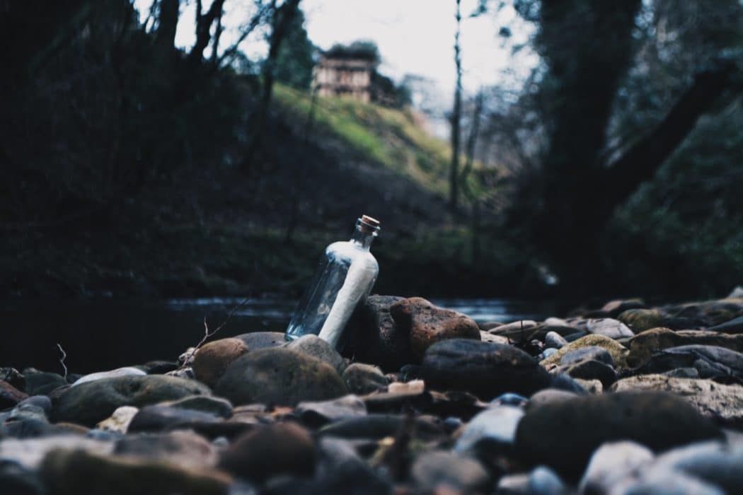 A close up of a message in a bottle on the rocks beside a river, a grassy hillside in the background.