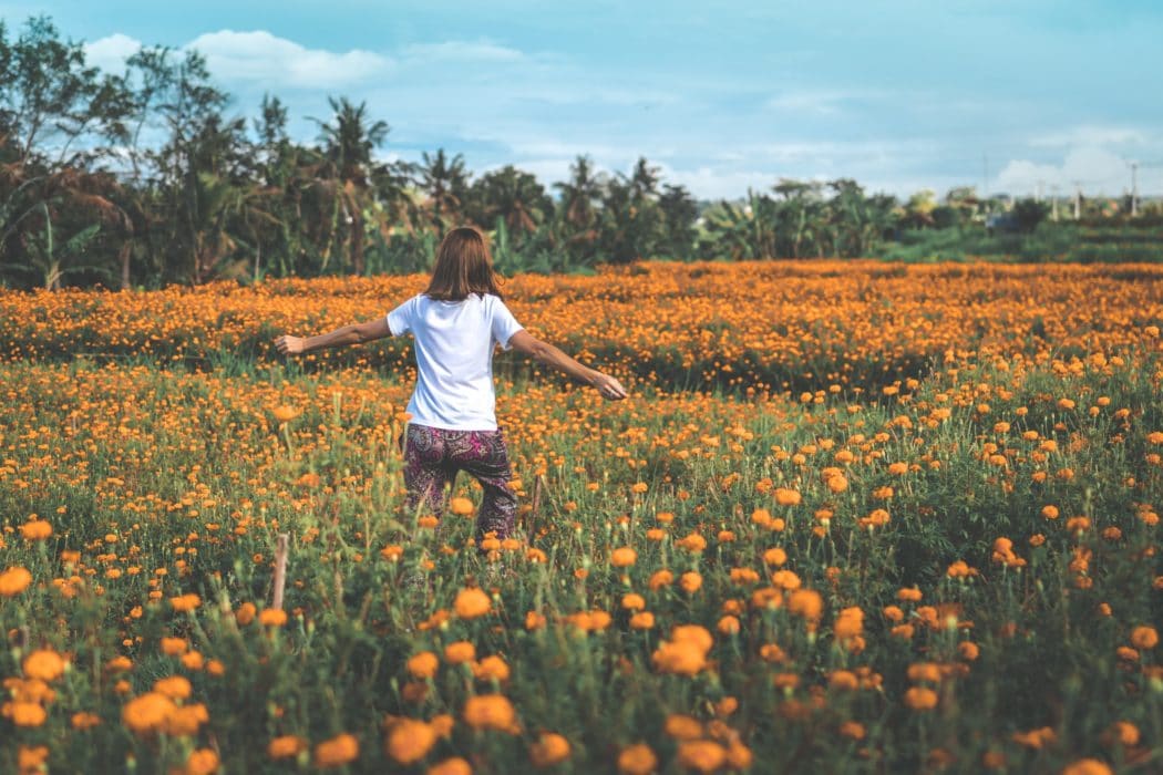 A young woman running through a meadow of flowers with palm trees lining the background.