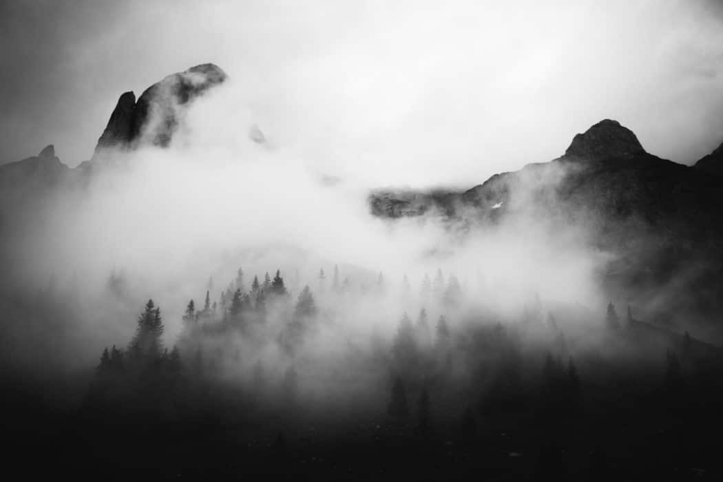 A mountain range with a dark, thickly wooded, forest below it, the whole scene shrouded in thick fog.