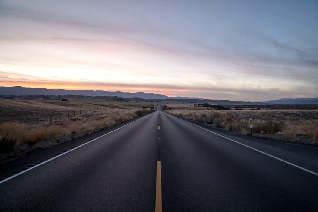A two lane road running through a grassy landscape, the sun setting over a mountain range on the horizon.
