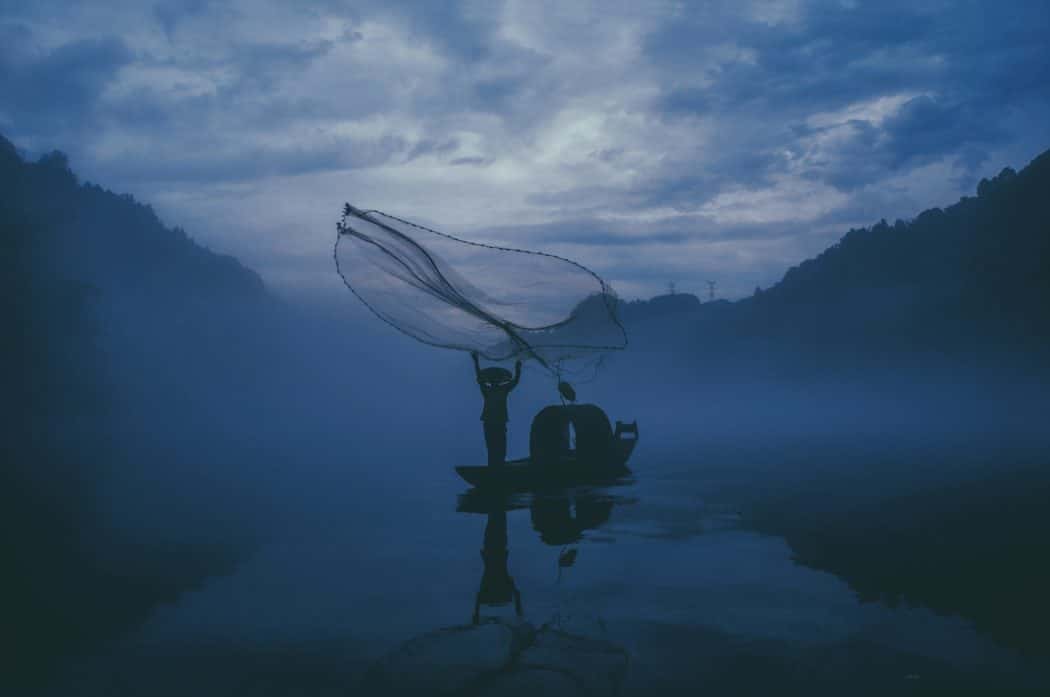 A fisherman casting out a net over a canoe in the middle of a dimly lit river, dark clouds hanging over head and trees lining either side of them. 