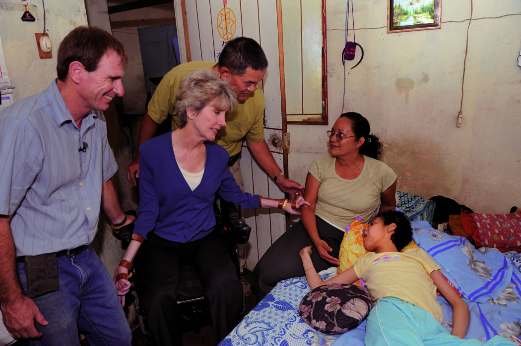 Joni and Ken inside a house in El Salvador, standing near a bedridden child who lies next to her mother; another man stands beside Joni