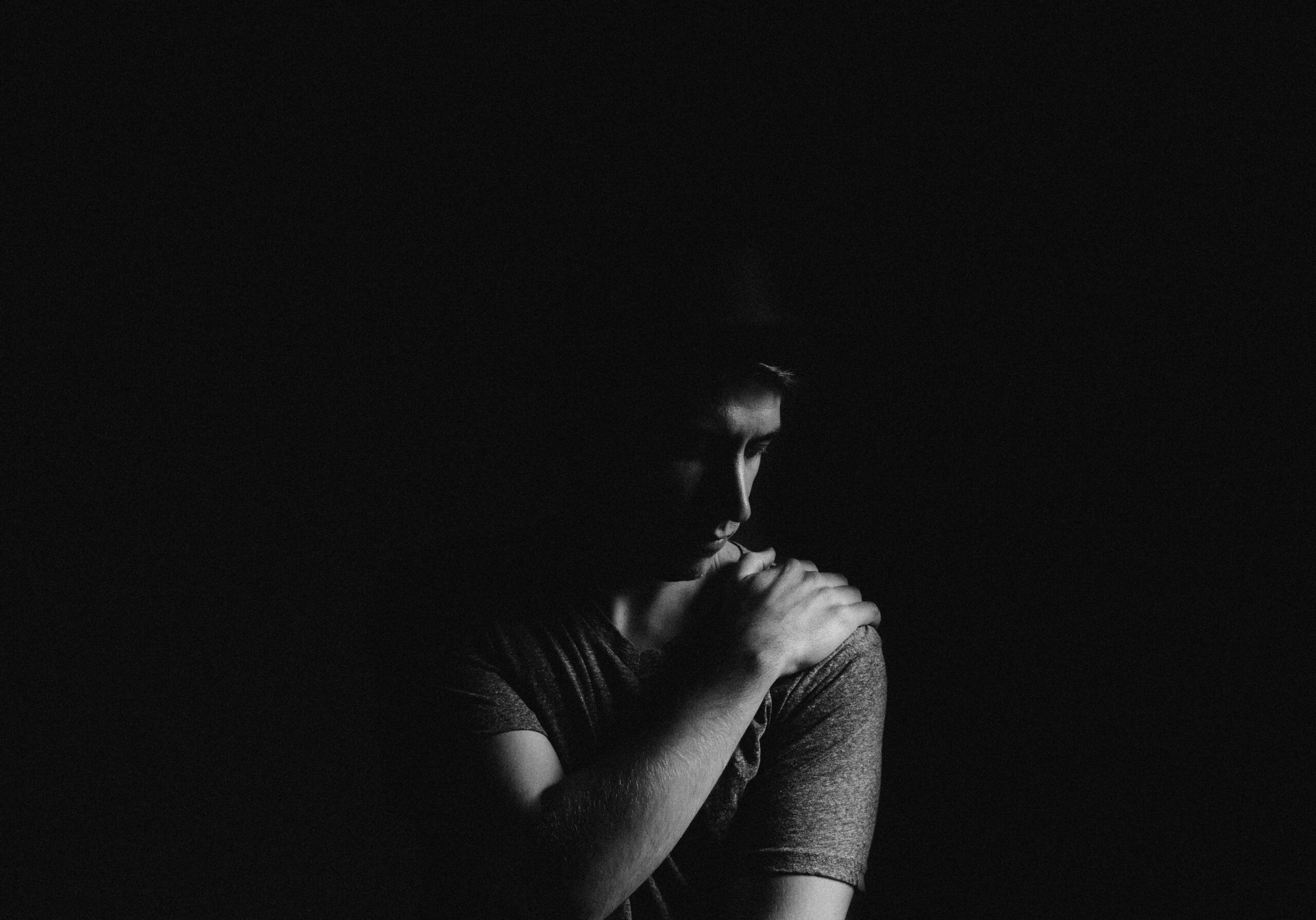 A black and white photo of a man in a dark room with his hand on his shoulder as he's looking down.