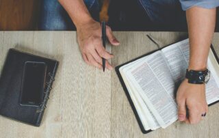 A picture of a man sitting at a desk with a bible open on the table and a journal with a pen in his hand and his phone on top of his journal.