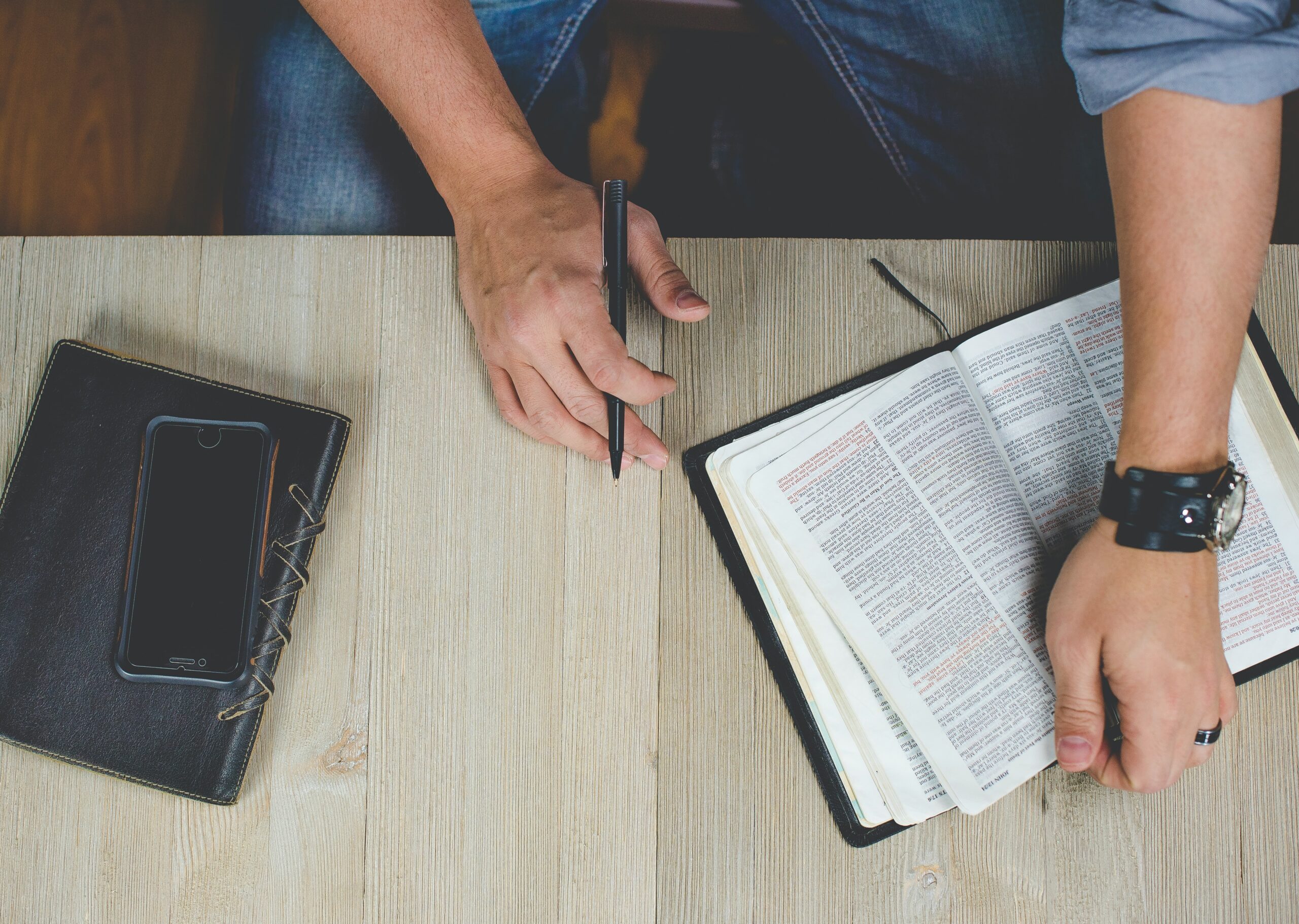 A picture of a man sitting at a desk with a bible open on the table and a journal with a pen in his hand and his phone on top of his journal.