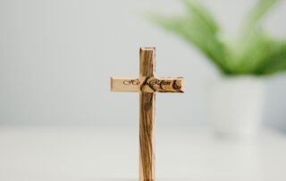 A picture of a miniature cross sitting on a white table with the words He is Risen in cursive on it.