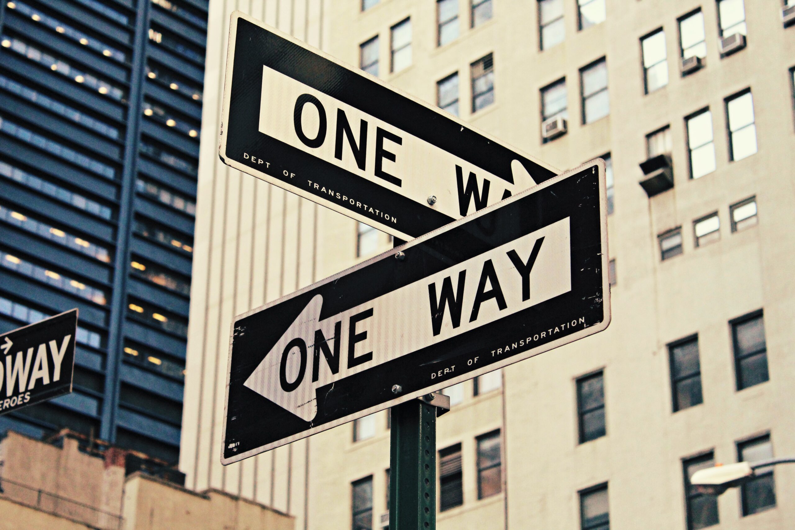 Close up of a pole with two signs going different directions saying "One Way" on them. Skyscrapers are behind them.