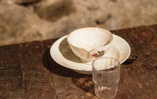 A broken teacup on a wooden table with a small clear glass cup in front of it.