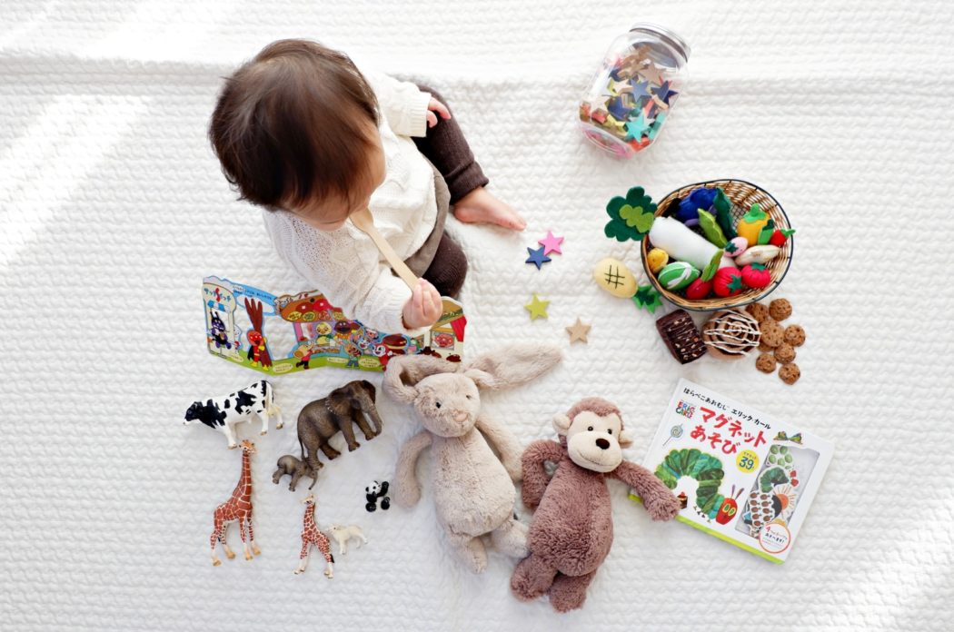 The view from above a baby playing on a white blanket putting a wooden spoon in his mouth with a bunch of colorful toys around him.