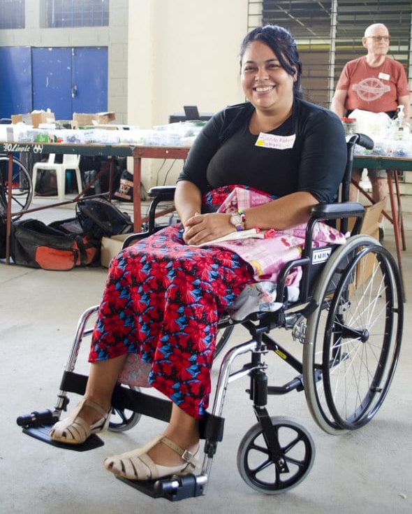 A woman seated in her wheelchair smiling at the camera with what looks like a work station with tools and supplies on it in the background.