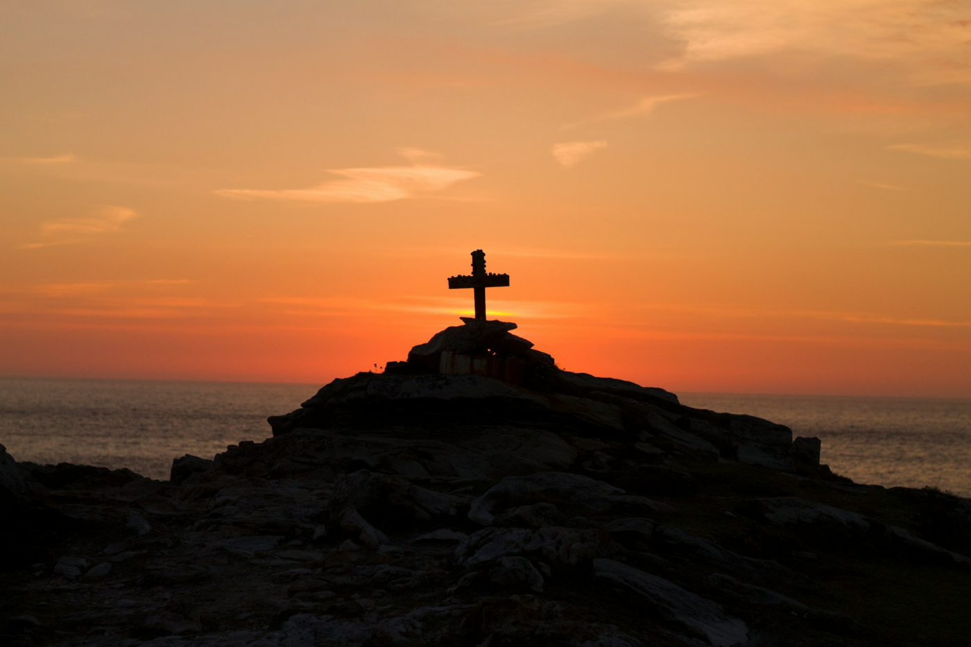 A cross silhouetted against a sunrise or sunset over a body of water, positioned on a rocky hill.
