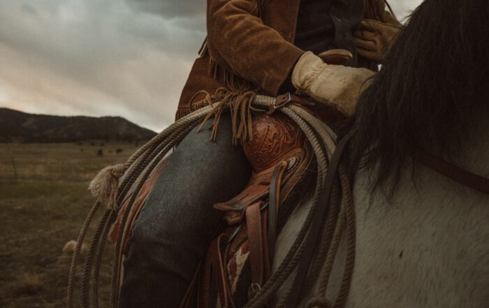 Close up of a cowboy riding a horse with a rope attached and a cloudy sky above a range behind him.