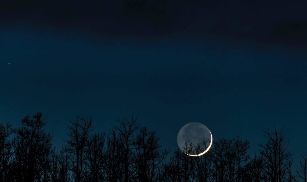 A beautiful photo of a deep, dark blue night sky with the moon rising over a line of silhouetted trees. You can see the entire outline of the moon, but the part that's shining is just a tiny sliver.