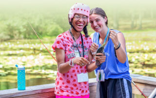 A photo of a young woman with a disability that requires her to wear a pink helmet along with her Joni and Friends buddy as they fish off of a dock.