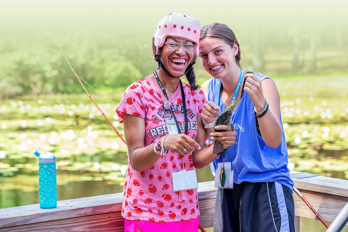 A photo of a young woman with a disability that requires her to wear a pink helmet along with her Joni and Friends buddy as they fish off of a dock.