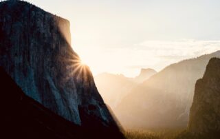A picture of the sun peaking around a cliff, lighting up the valley below and the mountain cliffs besides.