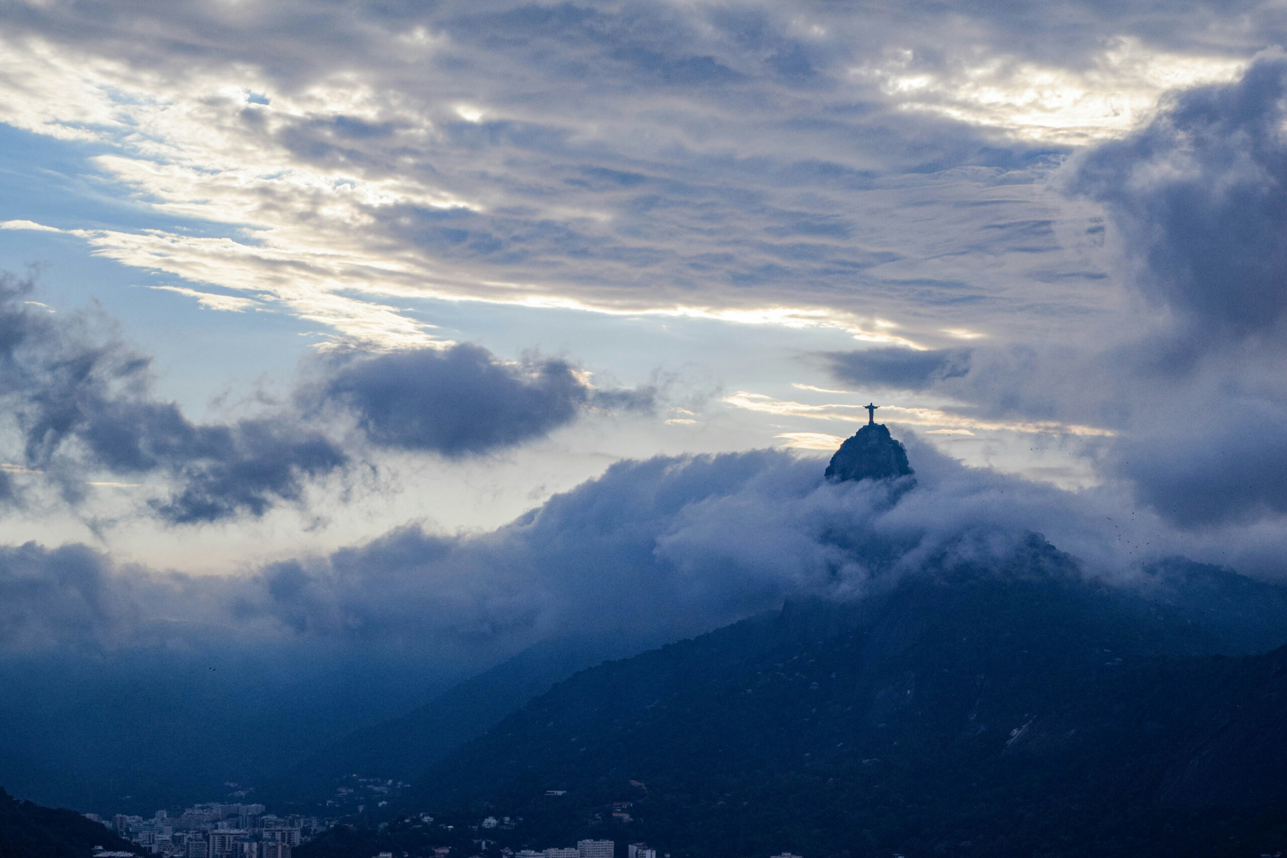 A picture of the Christ the Redeemer statue in Rio de Janeiro from afar with beautiful clouds moving all around it, the blue sky and sunlight peaking out.