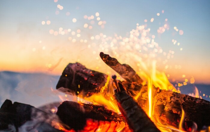 A close-up photo of logs on a fire with sparks flying up into the sunset sky.