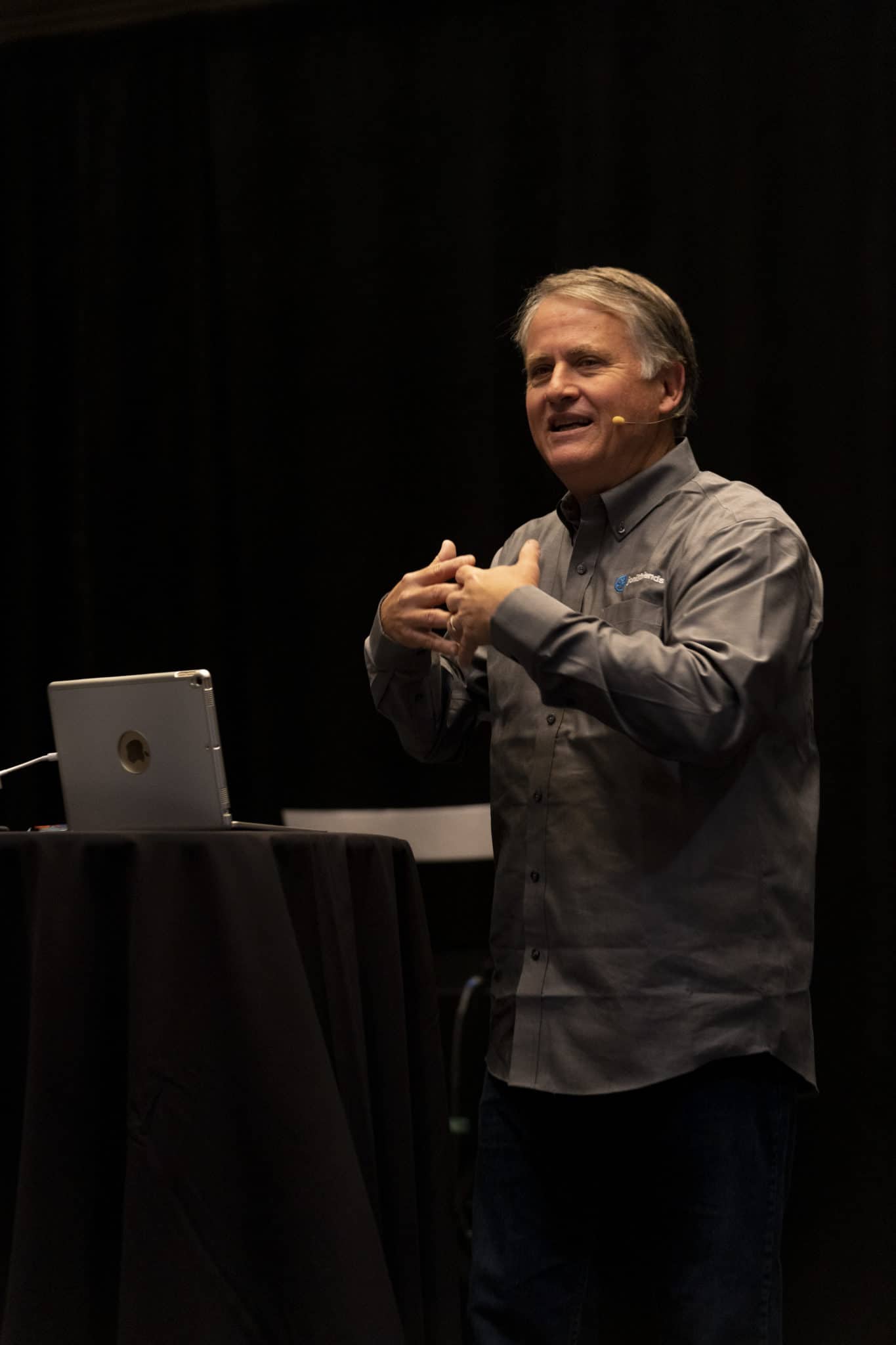 Randy Alcorn standing and speaking, with a laptop open in front of him. He is interlacing his fingers as he emphasizes a point, engaging with his audience while presenting.