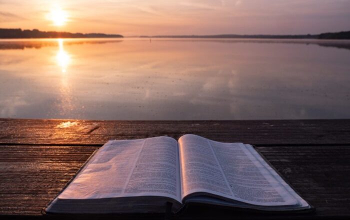 A Bible open on a wooden table facing a gorgeous body of water at sunset. The light is reflecting off the water in a pink glow.