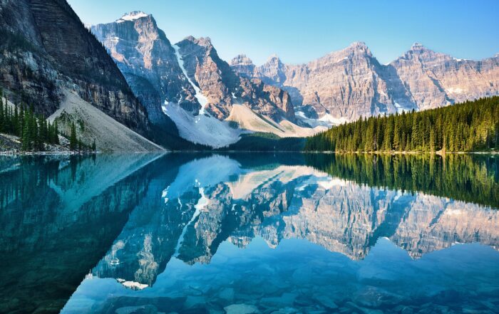 A picture of beautiful, rugged mountains towering over a crystal-clear lake with a forest of evergreen trees on the other side of the shore.