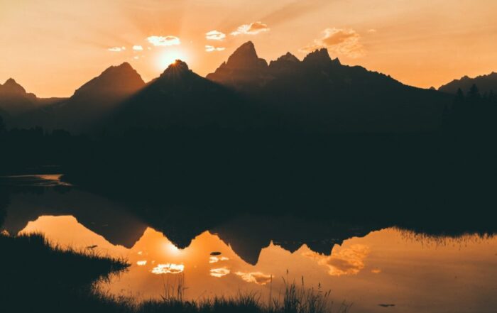 A picture of large mountain range silhouetted against a gorgeous orange sunset, a body of water reflecting it all below.