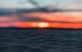 A close up of the sands on the beach with a bright orange sunset piercing the horizon in the distance.