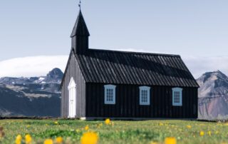 A church in a field with little yellow flowers around it and a mountain range behind it.