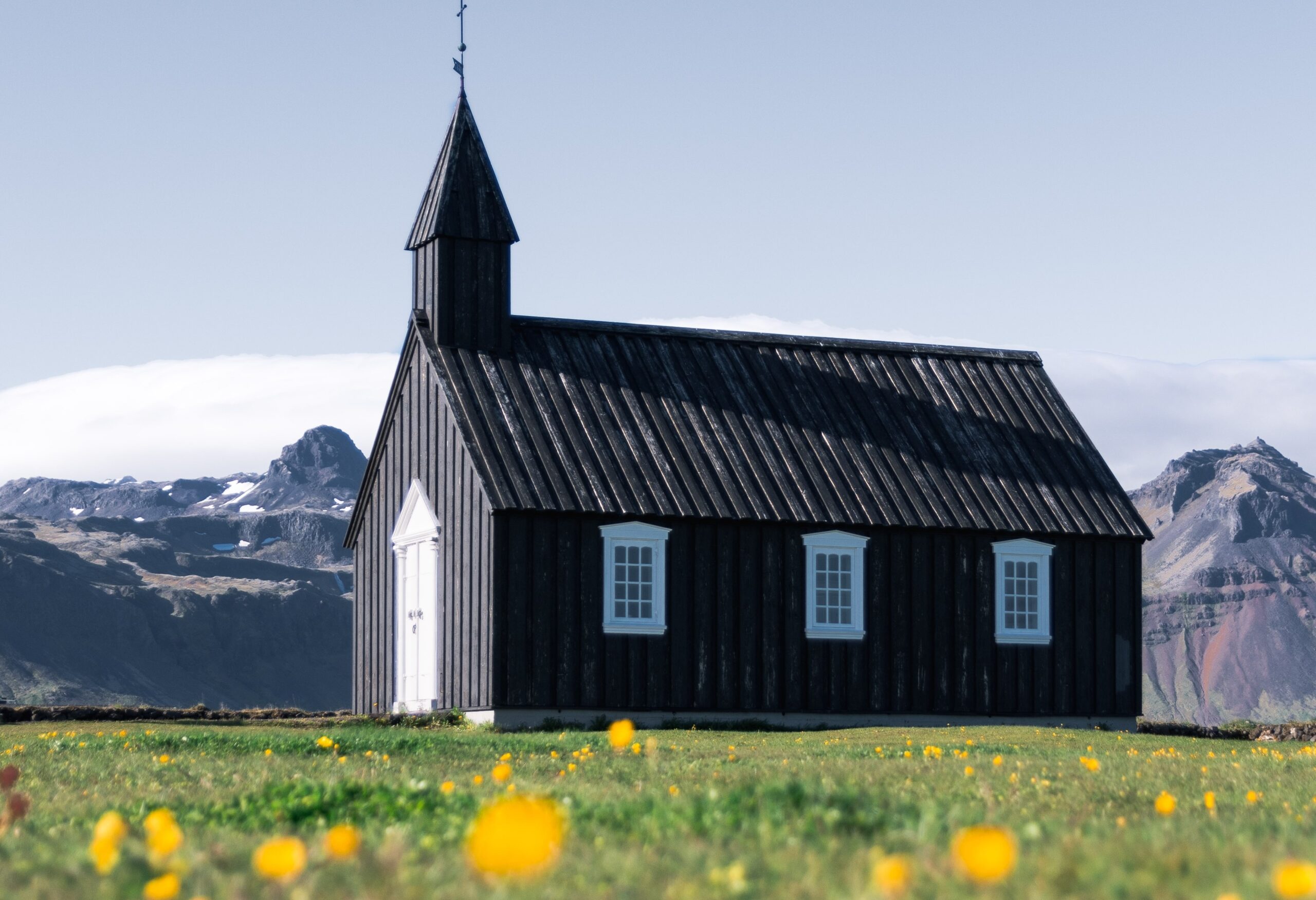 A church in a field with little yellow flowers around it and a mountain range behind it.