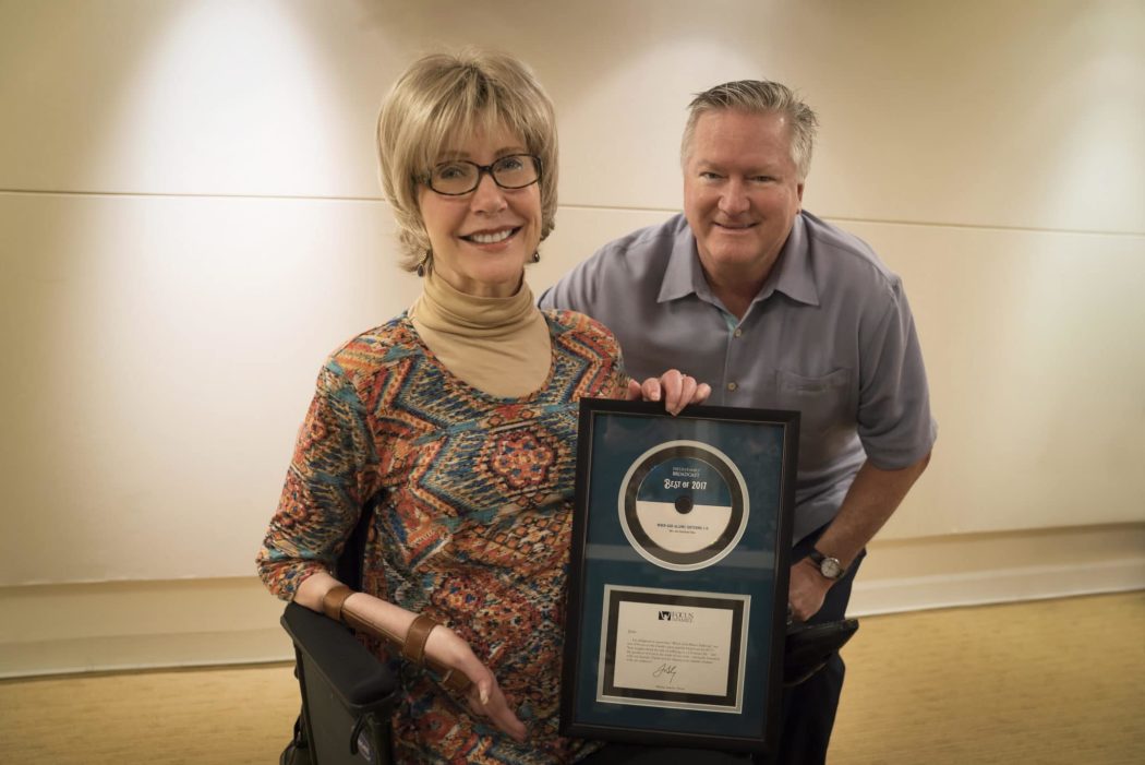Close up of Joni and Jim Sanders smiling at the camera as Joni holds up a framed record titled, "Best of 2017."