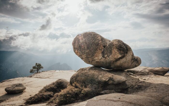 A picture of a boulder on top of a cliffside with the sun streaming through clouds above.