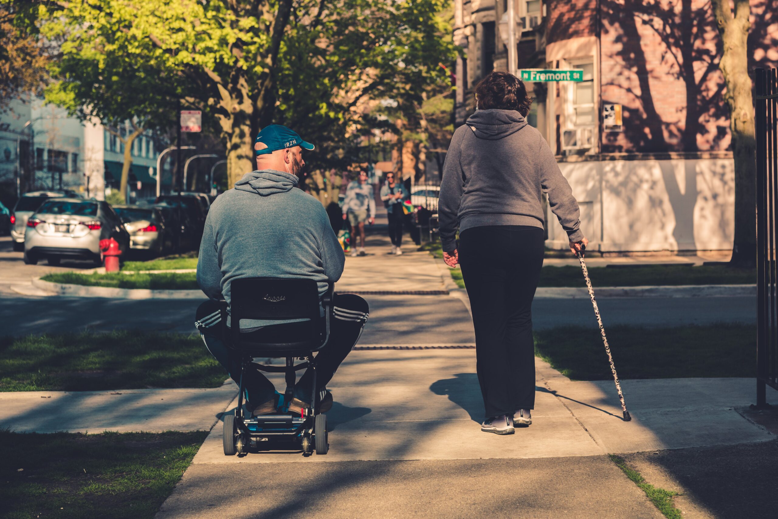 A man seated in an electric scooter beside a woman walking with a cane as they go down the sidewalk on a busy street.