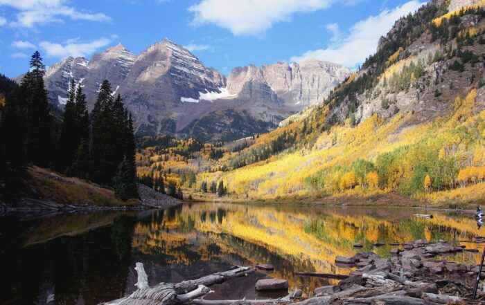 A view of a lake surrounded by beautiful rugged mountains and blue sky above and aspen trees covering the mountainside.