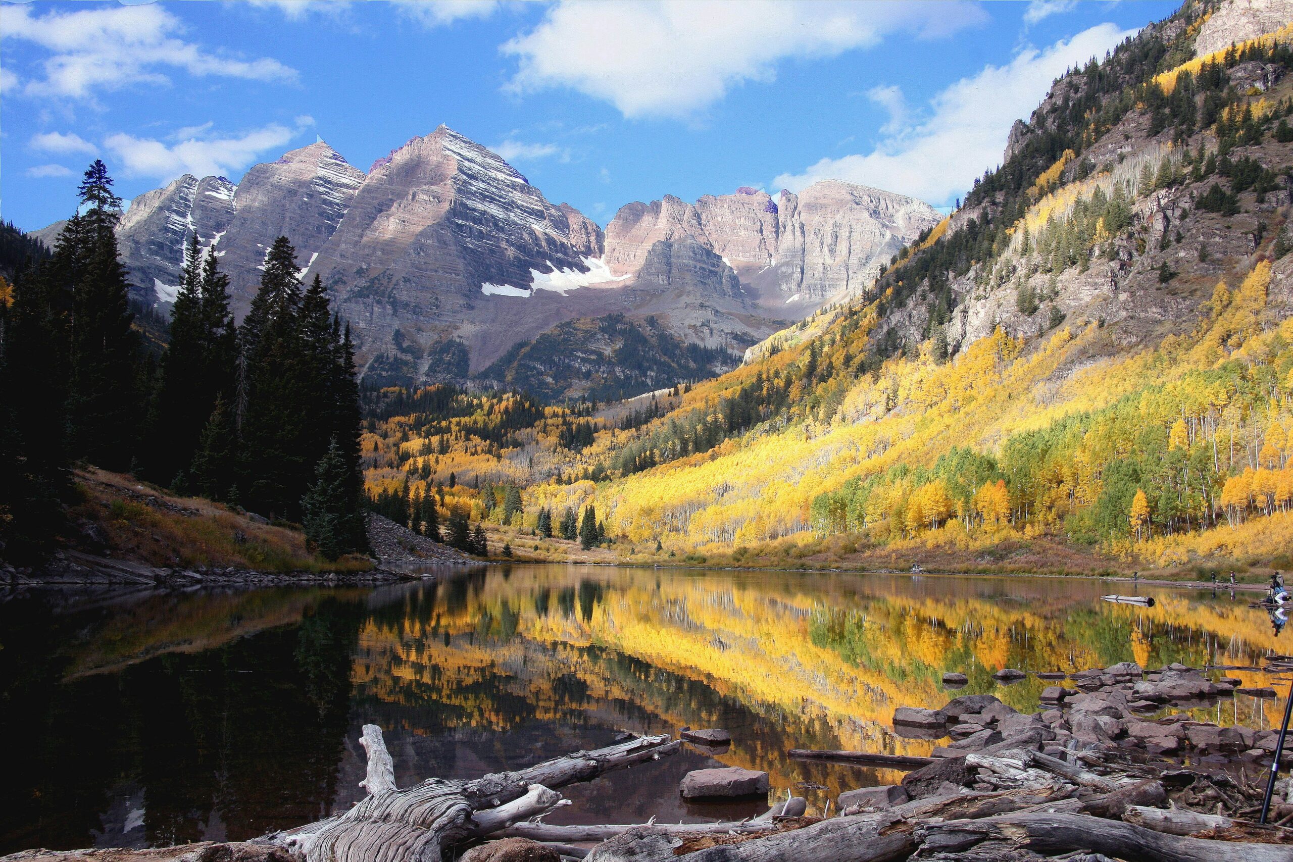 A view of a lake surrounded by beautiful rugged mountains and blue sky above and aspen trees covering the mountainside.