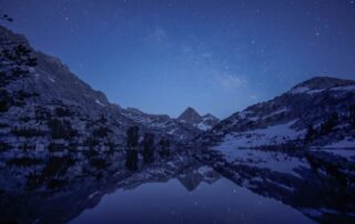 A view of snowy mountains over a body of water. The night sky above is covered in stars.