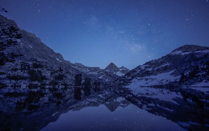 A view of snowy mountains over a body of water. The night sky above is covered in stars.