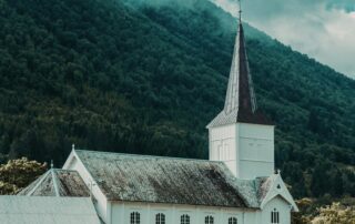 A picture of an old-fashioned white church building with a spire and a large mountain on the other side of it, covered in greenery.