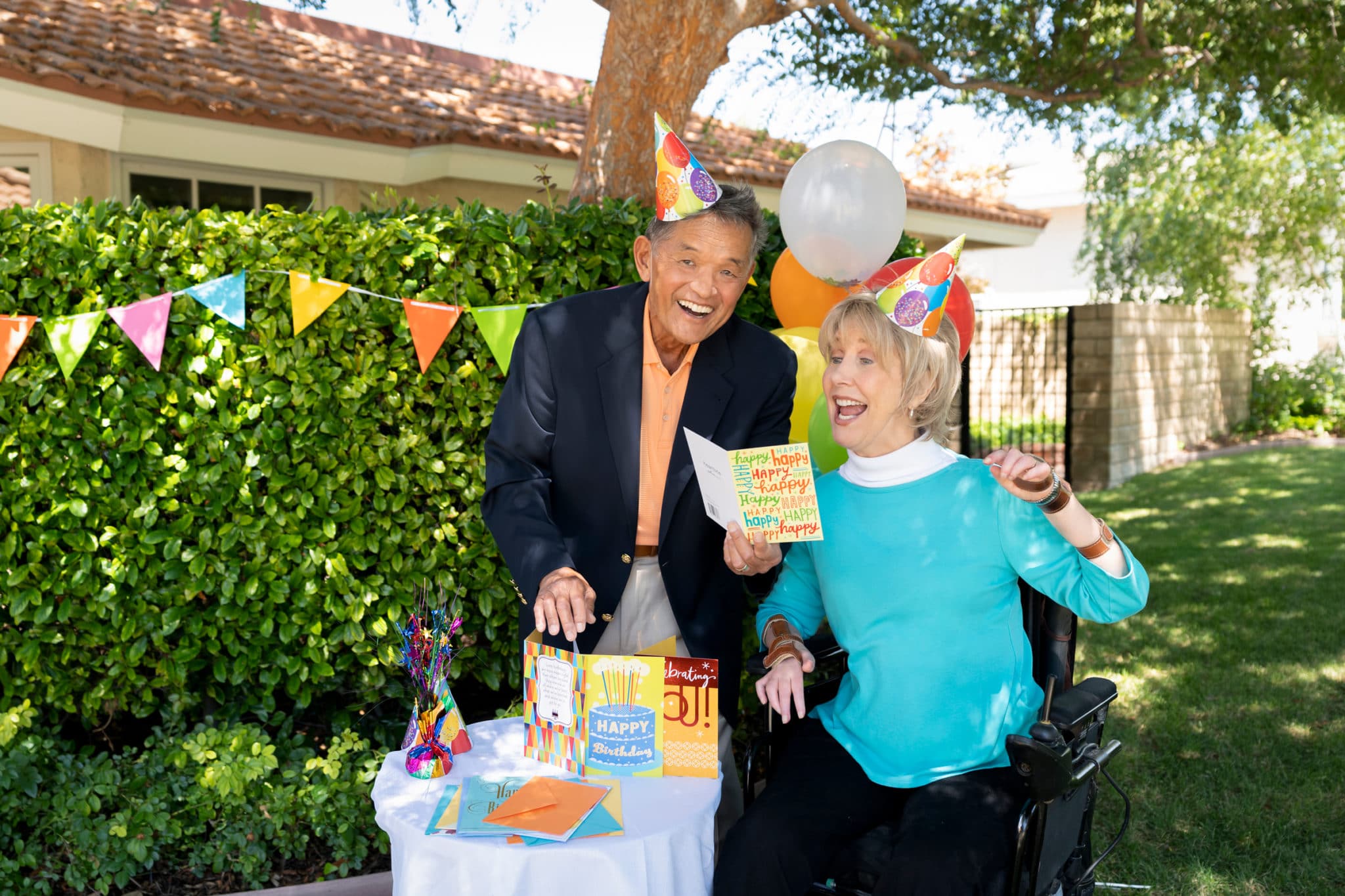 A scene of Ken standing next to Joni showing her a birthday card as they both smile wide, wearing party hats, surrounded by birthday decorations and balloons in what looks like a backyard.
