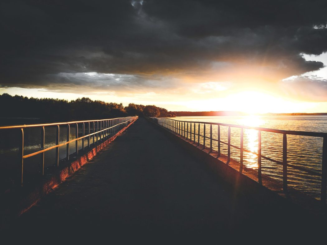 The view from the end of a bridge over a large body of water as the sun sets over an island in the background and reflects off of it the water.