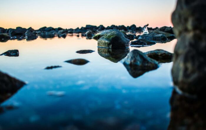 A close-up picture of a body of crystal clear water with some mossy rocks in it as the sun in setting in the background. The reflection of the rocks shining brightly in the water.