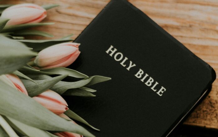 A picture of a Bible on a wooden table that says "Holy Bible" on the front and a bouquet of flowers laying on top of it.