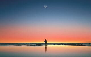 A silhouette of a man standing on a beach as the sun is setting into a deep pink color, the moon in the dark blue sky above.