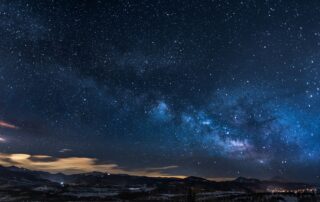 A picture of a starry sky full of thousands of bright stars and visible galaxies. Beneath is a small mountain range with snow at the peaks.