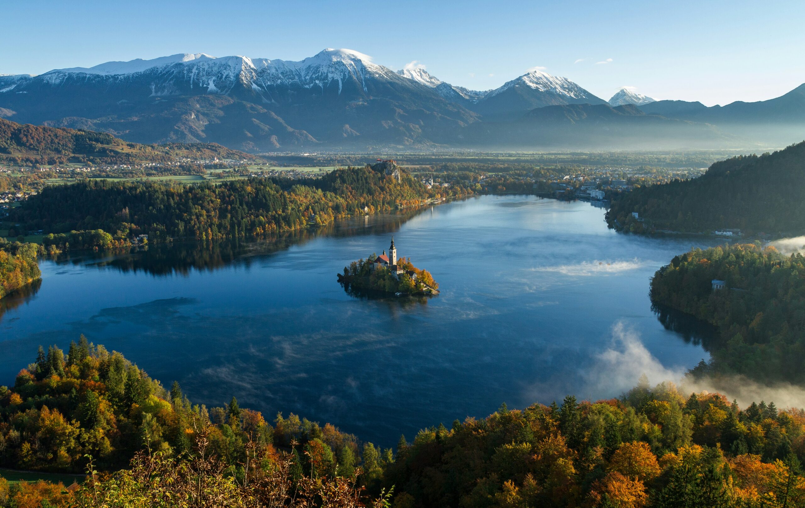 A picture of a mountain ranger with snow on top of it, below the mountain is a large lake and a landscape of orange and green trees.