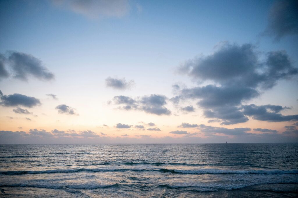 A picture from the shore of the beach as the sun it setting over the ocean waters. There are speckled clouds in the sky.